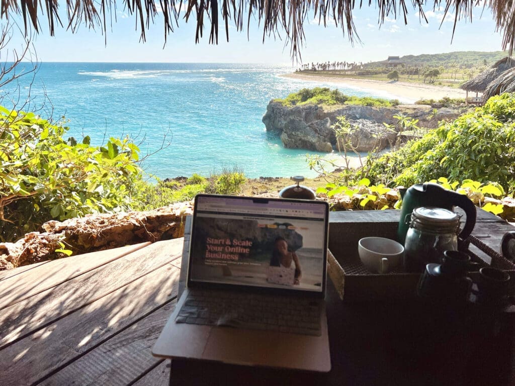 Laptop open to The Wanderlover Website on a wooden balcony overlooking bright blue waters and green fauna in Rote, Indonesia.